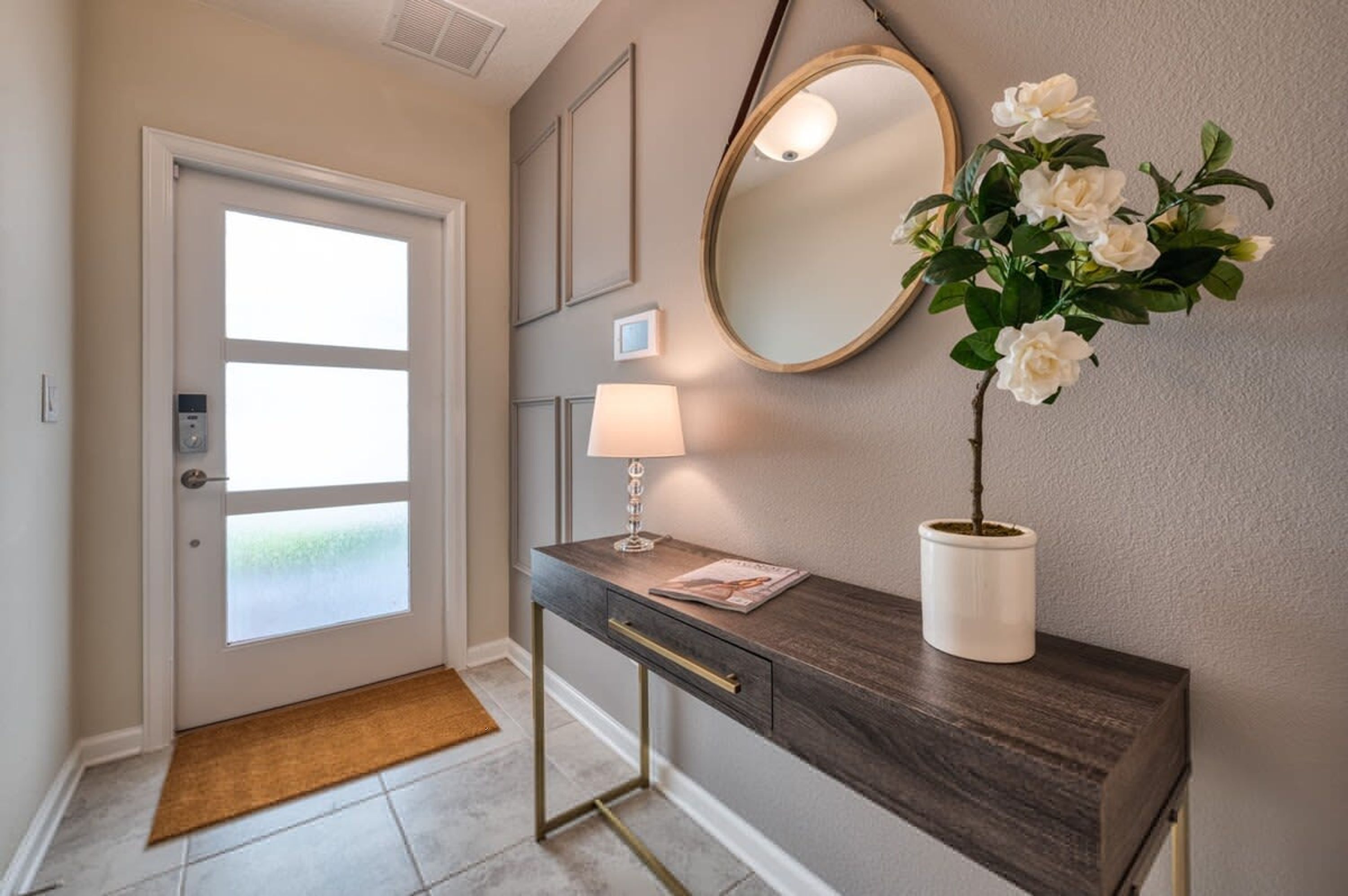 A hallway with a door, a desk with a lamp, a mirror, and a potted plant.