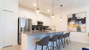 A modern kitchen with white cabinets, a stainless steel refrigerator, a microwave, and a bar area with bar stools. - Chic Modern Marvel-spacious Themed Bedrooms Private Pool (Kissimmee)