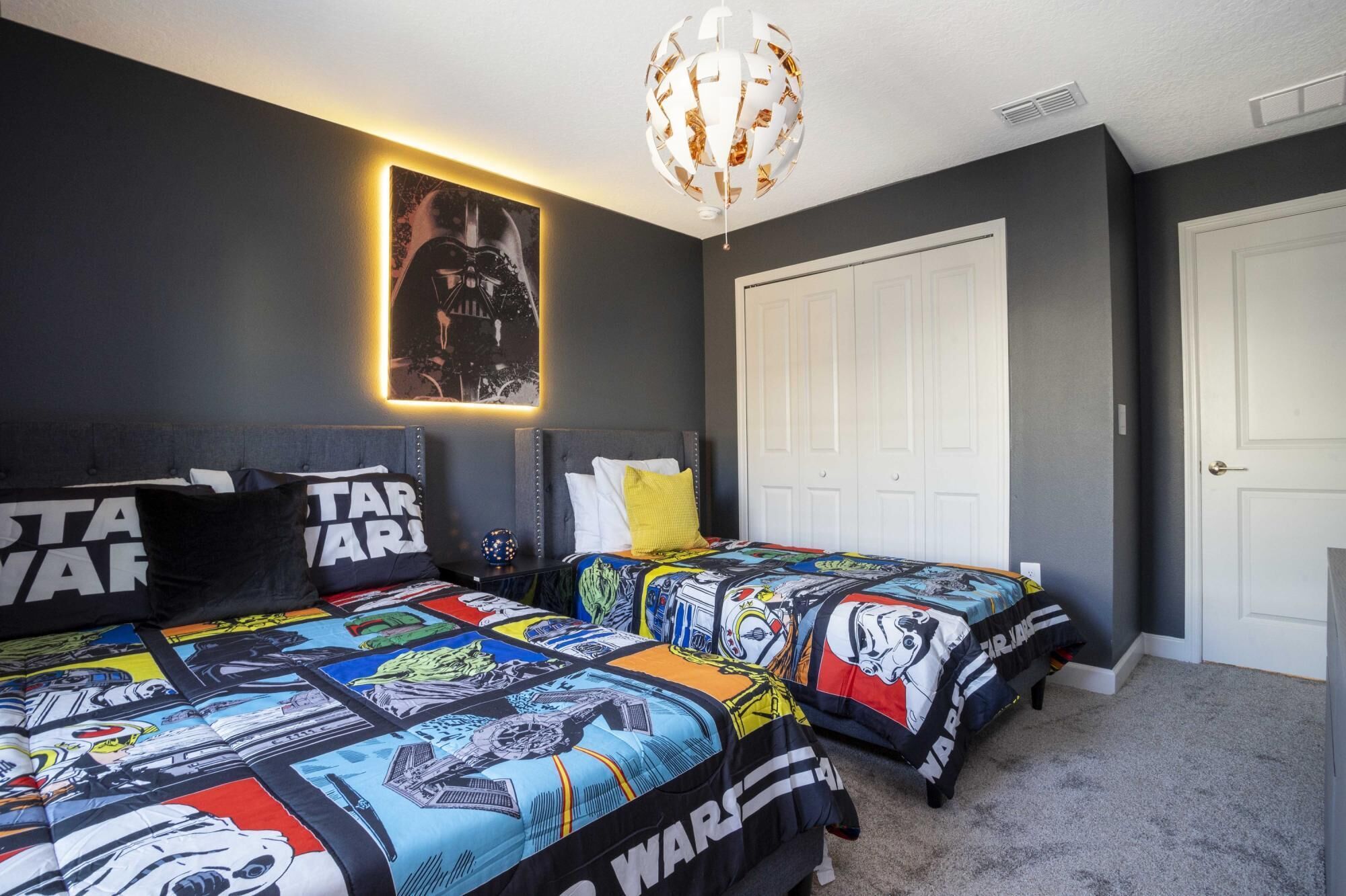 A bedroom with two beds, Star Wars themed bedding, a Darth Vader poster, and a modern chandelier.