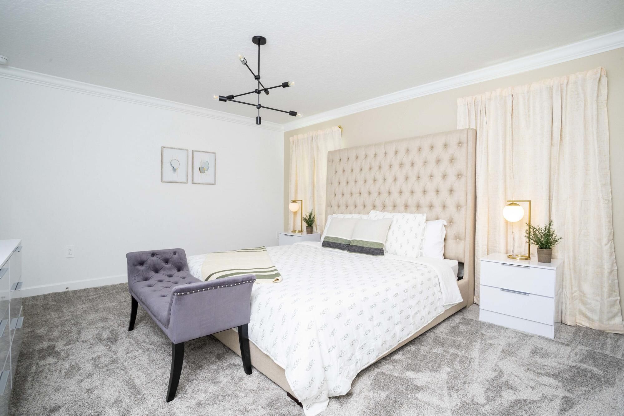 Ein Schlafzimmer mit einer gesteppten Kopfstütze, einem grauen gesteppten Sessel, einer weißen Bettdecke mit Muster und einer modernen Pendelleuchte.