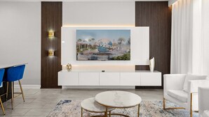 Ein modernes Wohnzimmer mit großem Flachbildfernseher an einem weißen Schrank, einem blauen Sessel, einem weißen Sofa und einem runden Couchtisch.