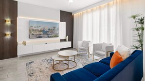Ein modernes Wohnzimmer mit einem blauen Sofa, weißen Sesseln, einem runden Couchtisch und einem großen, an der Wand montierten Fernseher.