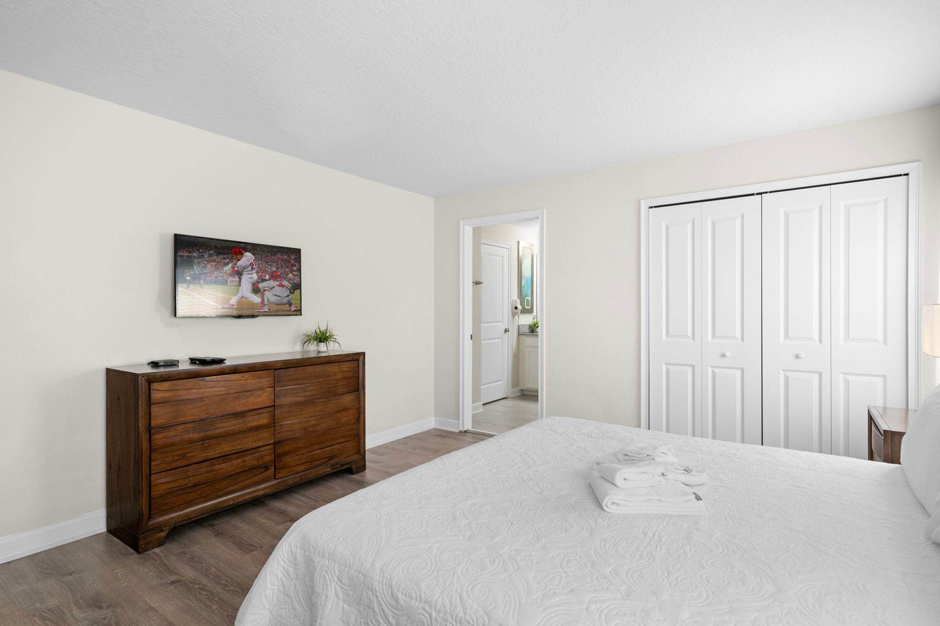A bedroom with a large bed, a wooden dresser, a painting on the wall, and a white door.