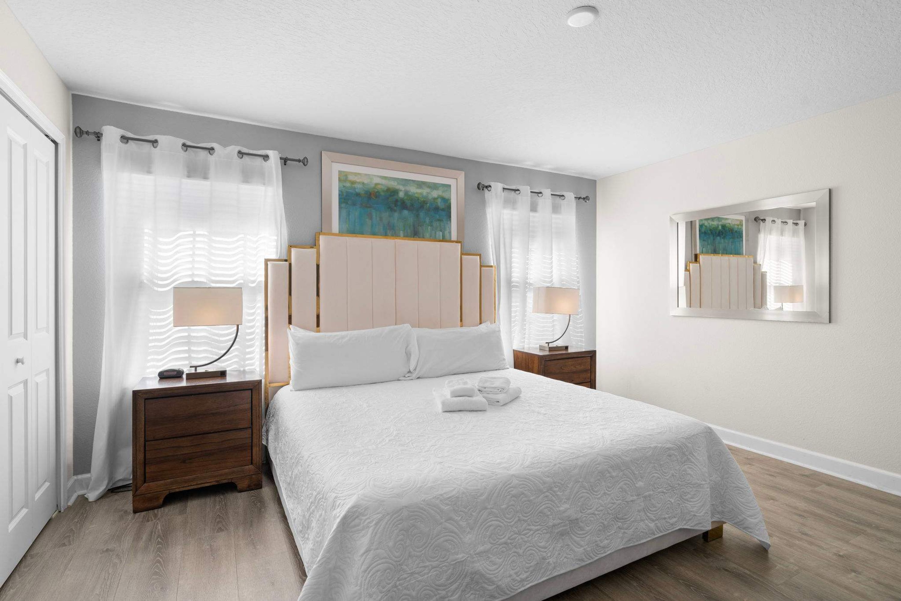 A bedroom with a large bed, two bedside tables, a nightstand, a painting on the wall, and a mirror.