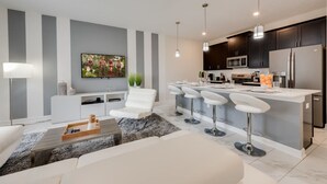 A modern kitchen with dark cabinets, a central island with bar stools, a flat-screen TV, and a dining area with a white sofa and a coffee table. - Cozy Home Prime Disney Parks Location (Kissimmee)