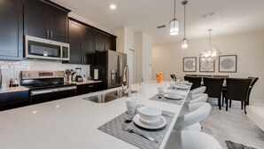 A modern kitchen with dark cabinets, stainless steel appliances, and a central island with seating. - Cozy Home Prime Disney Parks Location (Kissimmee)