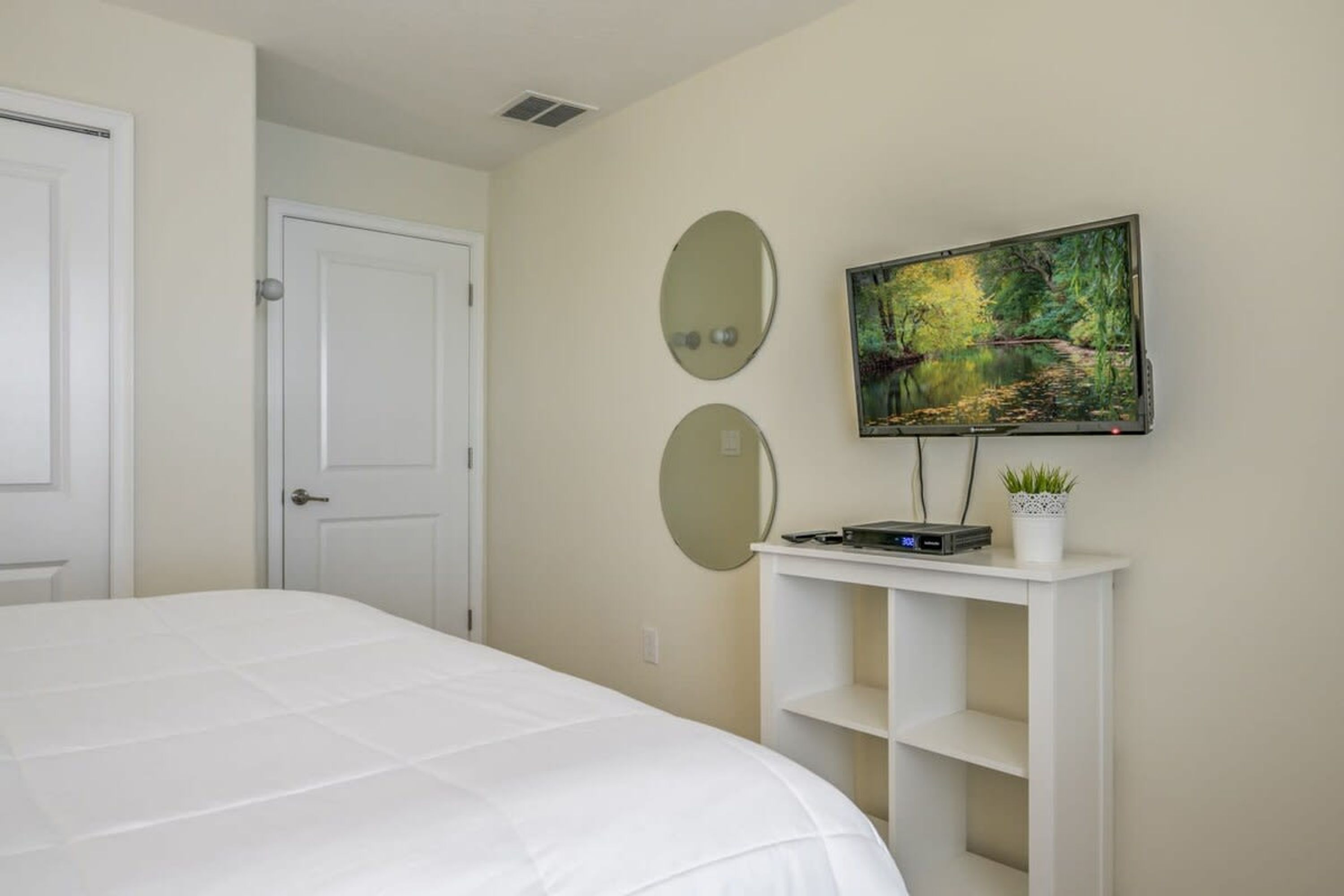 A bedroom with a bed, a TV mounted on the wall, and a small white console table with a plant.