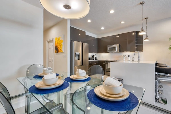 A modern kitchen with stainless steel appliances, a dining area with glass tables and blue placemats, and a yellow artwork on the wall.