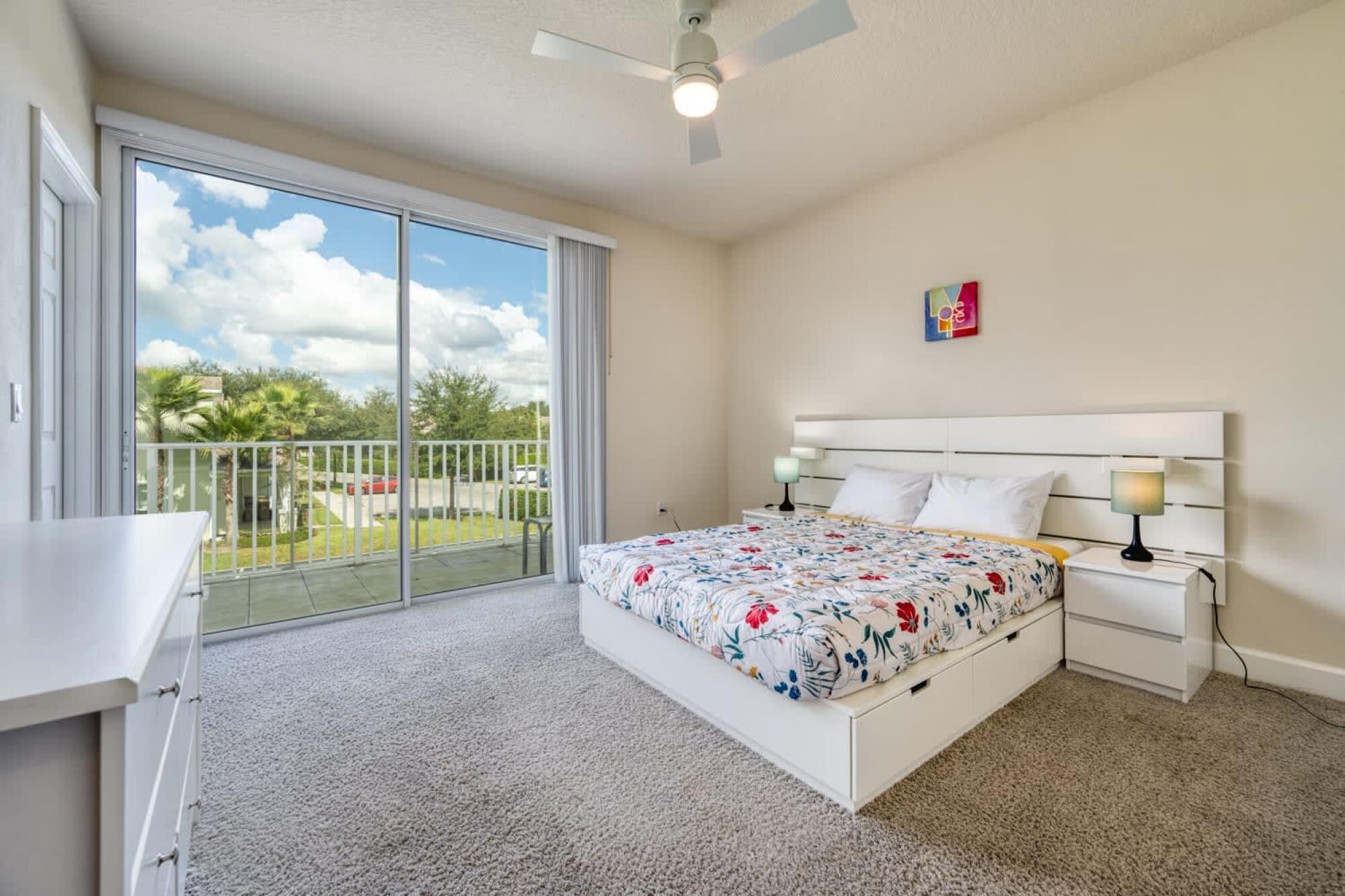 A bedroom with a large bed, a ceiling fan, a nightstand with a lamp, and a view of a balcony with a railing.