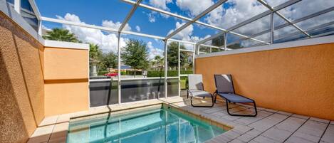 A covered outdoor swimming pool with a glass roof, two lounge chairs, and a view of the neighborhood.