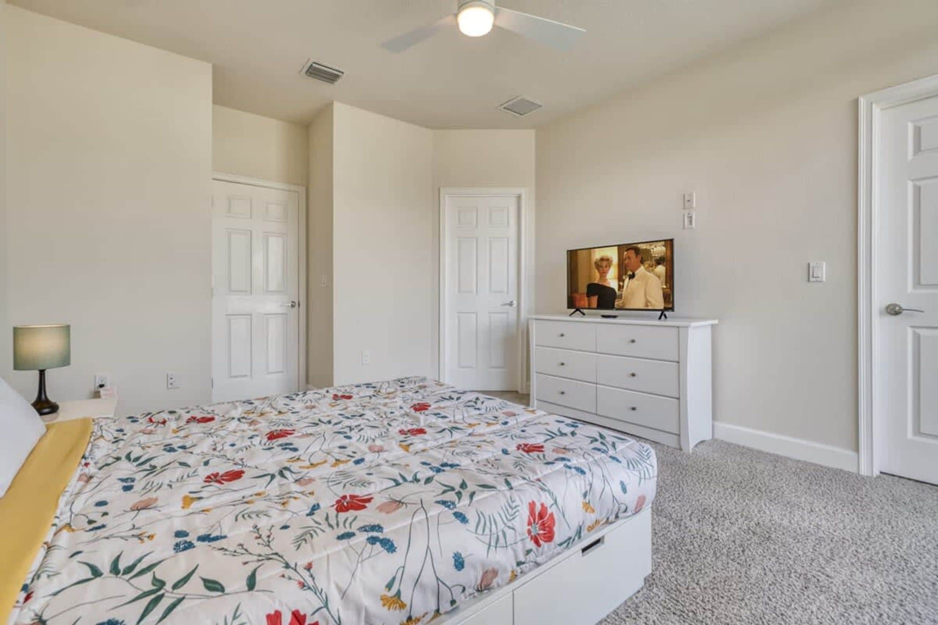 A bedroom with a floral bedspread, a white dresser, a television on a stand, and a ceiling fan.