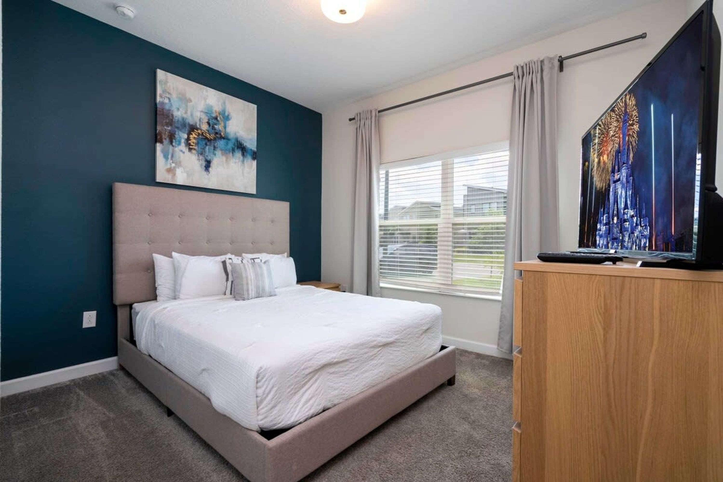 A bedroom with a bed, a TV, a painting, and a window with curtains.