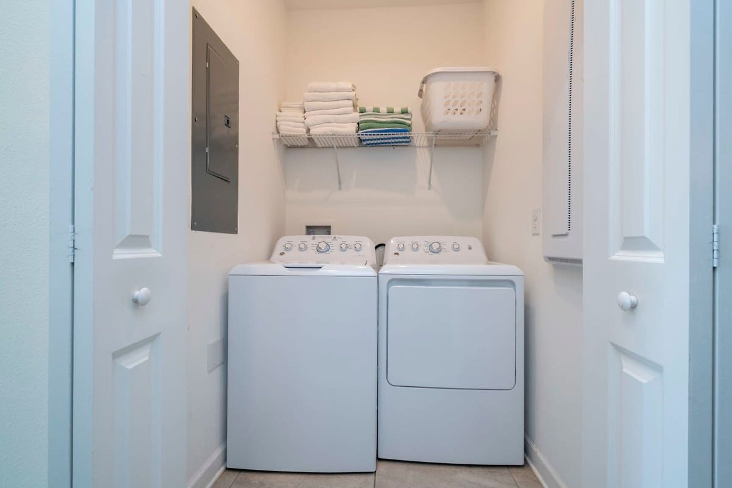 A laundry room with two white front-loading washing machines and a shelf above holding folded towels and a laundry basket.