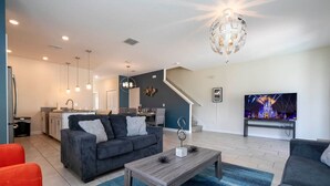 A modern living room with a grey sofa, a wooden coffee table, and a TV. A kitchen is visible in the background with a staircase leading to another level. - Cozy Getaways - Spacious Ideal Location (Davenport)