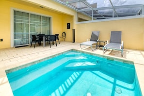 Ein Swimmingpool mit Liegestühlen und einem Tisch, umgeben von einem gelben Gebäude mit Glastür und einer überdachten Terrasse.