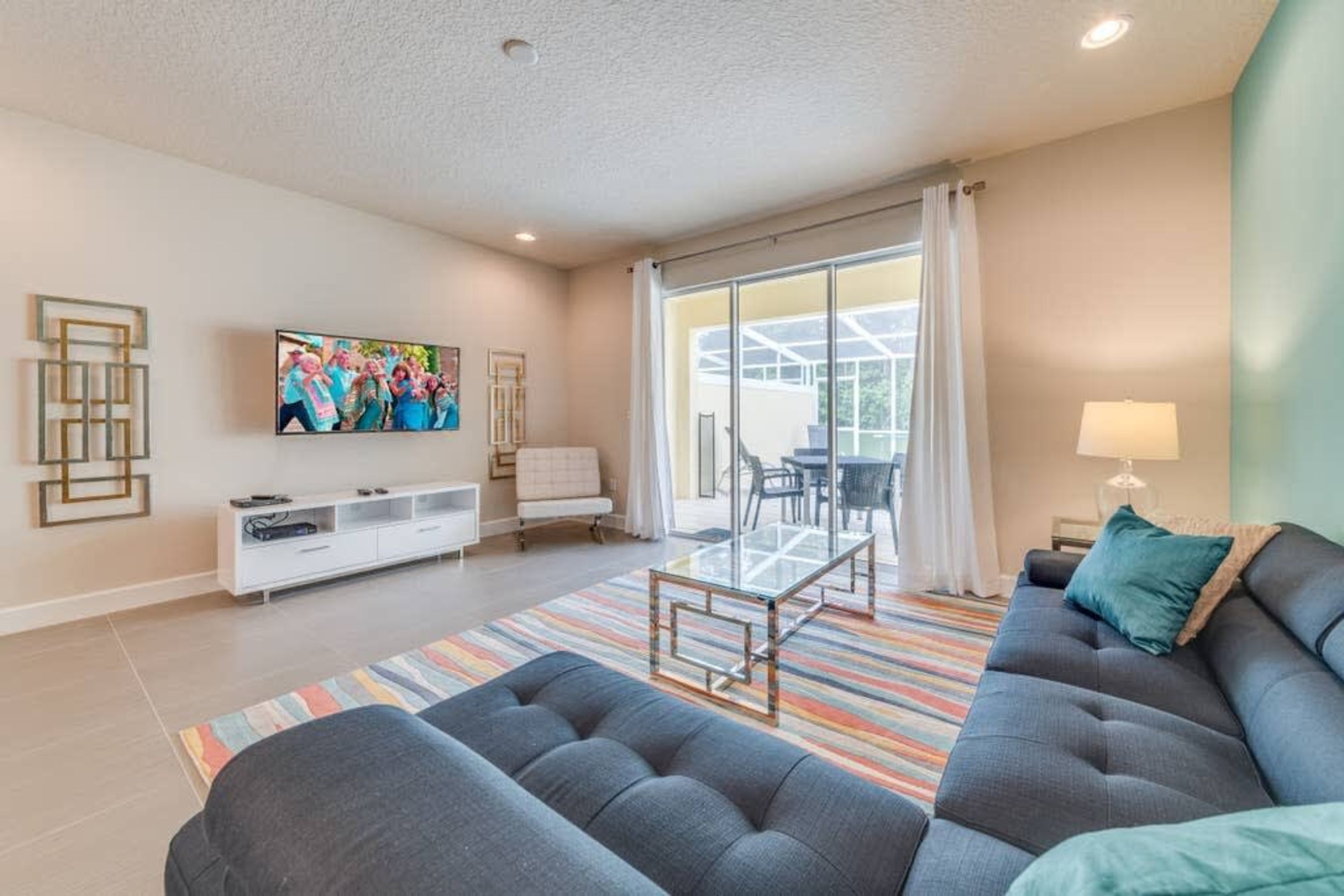 A modern living room with a grey sofa, a glass coffee table, and a colorful striped rug. There is a flat-screen TV mounted on the wall, a white entertainment unit, and a large window with curtains.