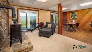 A cozy living room with a stone wall, a black wood-burning stove, a grey sofa, and a dining area with a wooden wall and a window. - Sparkling Bear Lodge | Epic views! | Touch the stars (Mount Jackson)