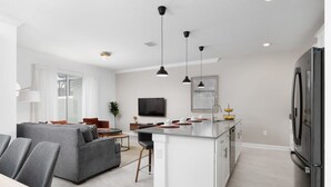 Una cocina moderna con una isla central, un comedor con sillas tapizadas en gris, y una sala de estar con un sofá gris.