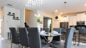 A modern dining area with a glass table and black chairs, a chandelier, and a kitchen in the background. - Modern Marvel-spacious Themed Fun-filled Family Getaway (Kissimmee)