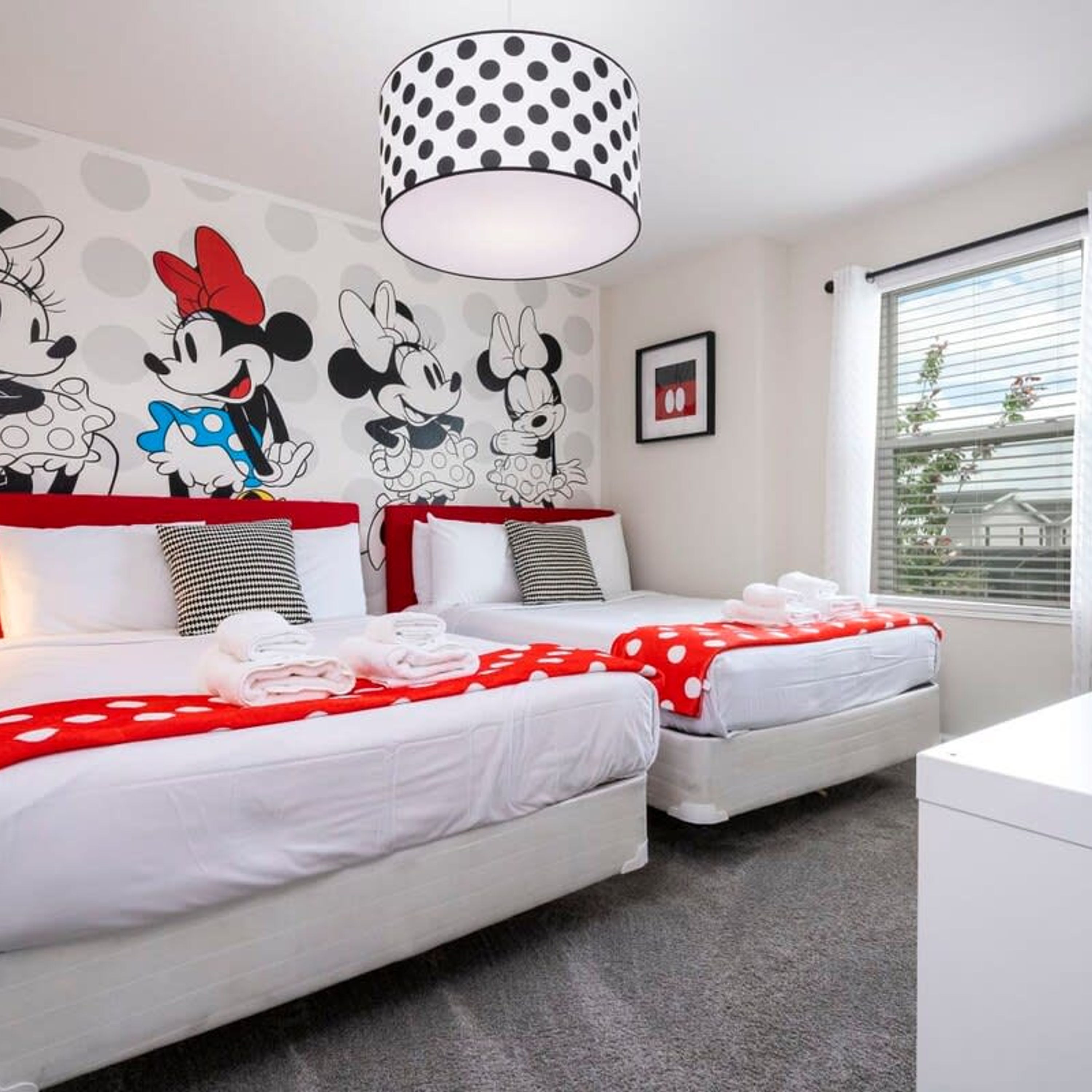 A hotel room with two beds, a Mickey Mouse mural, a polka dot lamp, and a window with blinds.