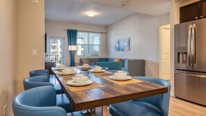 Une cuisine moderne avec un réfrigérateur en acier inoxydable, un coin repas avec une table en bois et des chaises bleues, et un salon avec un canapé et une fenêtre équipée de stores.