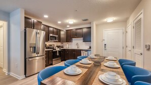 Une cuisine moderne avec des meubles de couleur sombre, des appareils en acier inoxydable et des chaises bleues disposées autour d’une table à manger en bois, préparée pour accueillir quatre personnes.