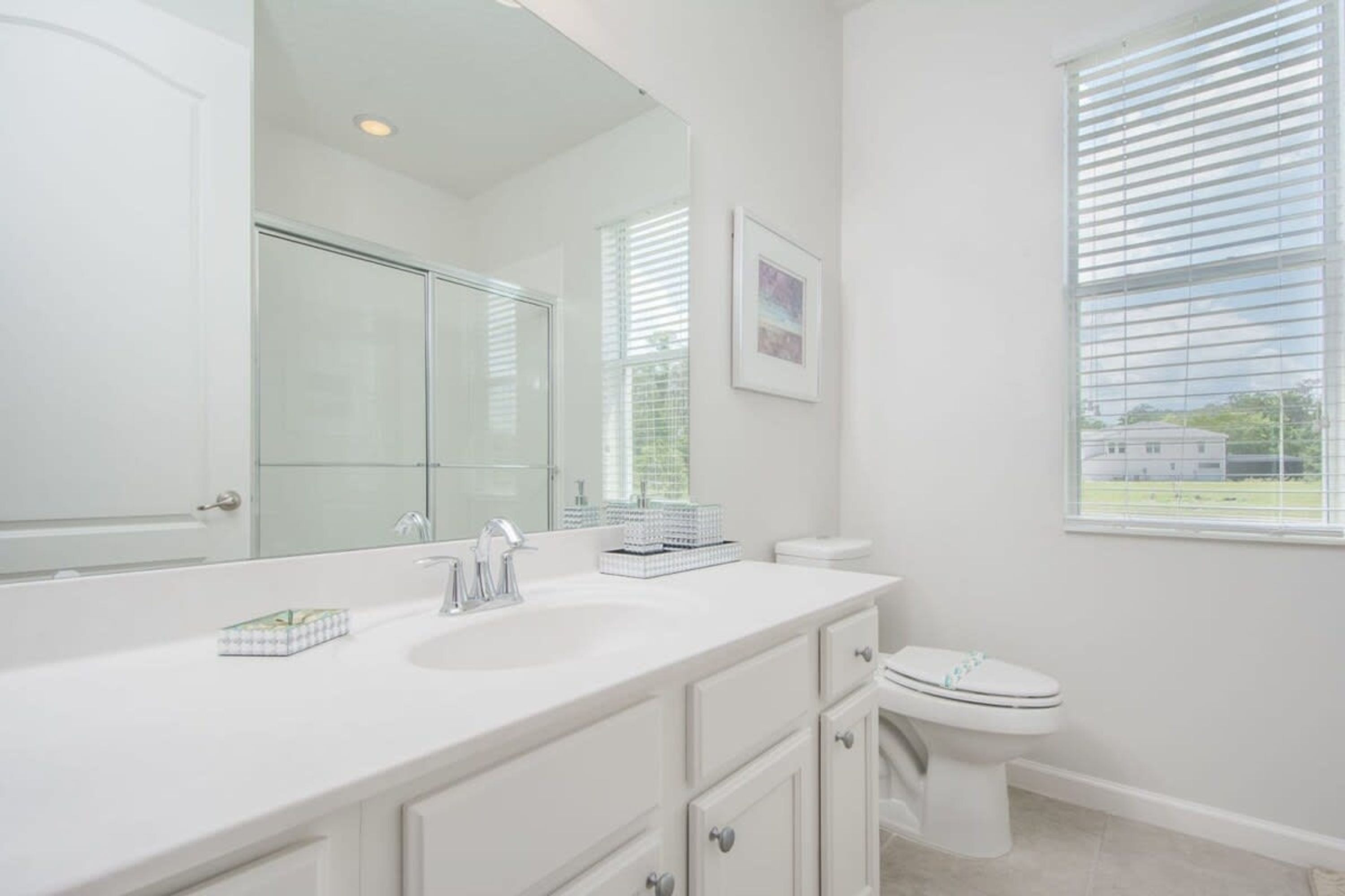 A bathroom with a white vanity, a large mirror, a toilet, and a window with blinds.