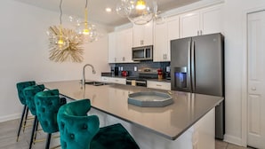 Une cuisine moderne avec des meubles blancs, un grand îlot central, des chaises de couleur turquoise et un lustre sphérique original.