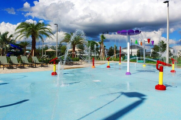 A water park with multiple water features, including fountains and water slides, surrounded by palm trees and lounge chairs.