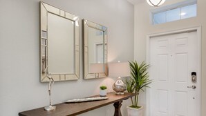 Een hal met een houten console, twee wandspiegels, een kleine potplant, een lamp en een witte deur met deurmat.