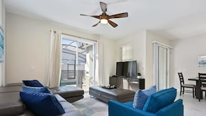A modern living room with a sofa, ottoman, and a blue armchair. There is a ceiling fan, a flat-screen TV, and a dining area with chairs.