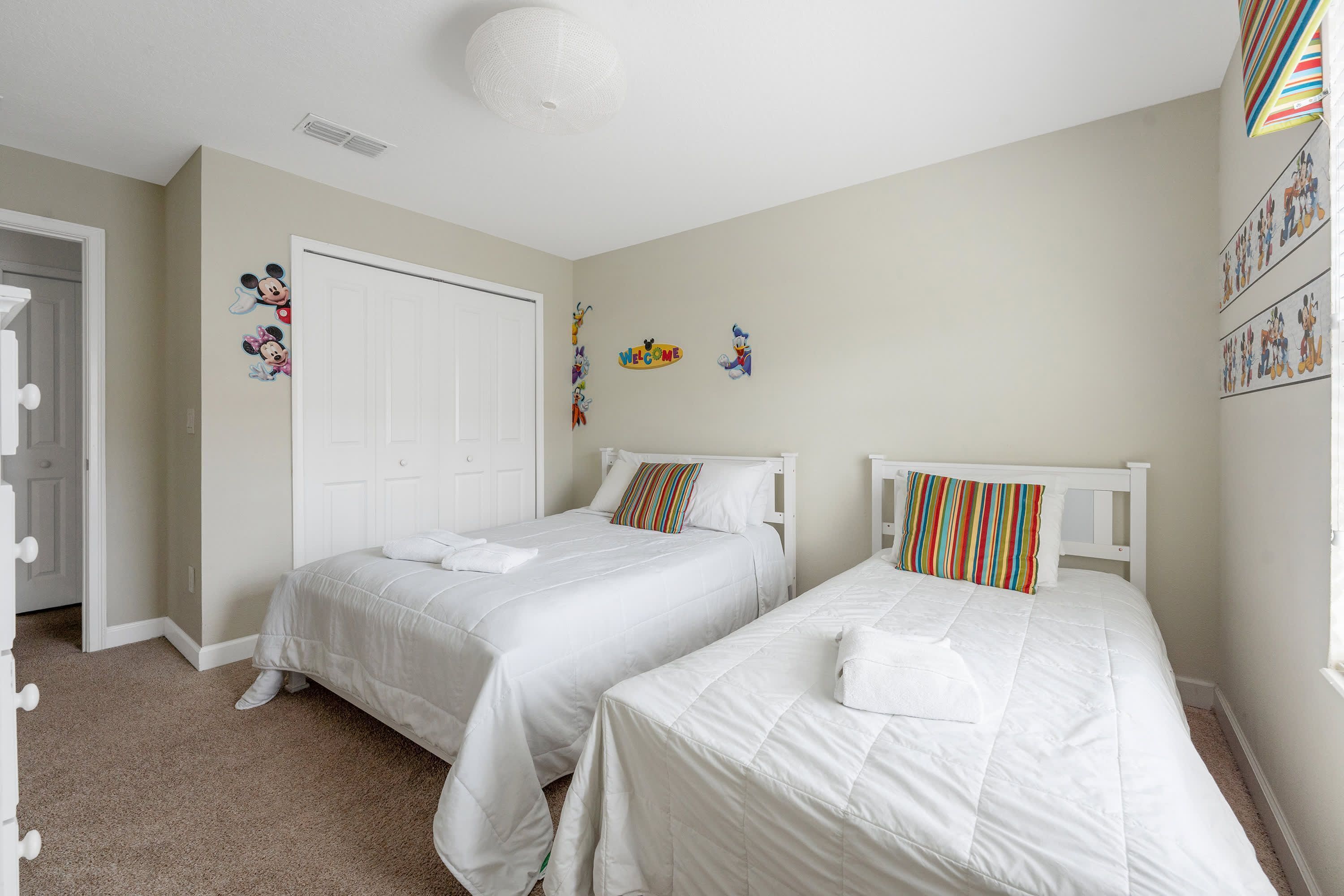 Quarto com duas camas, roupa de cama branca, almofada listrada e decorações nas paredes incluindo personagens de desenhos animados.