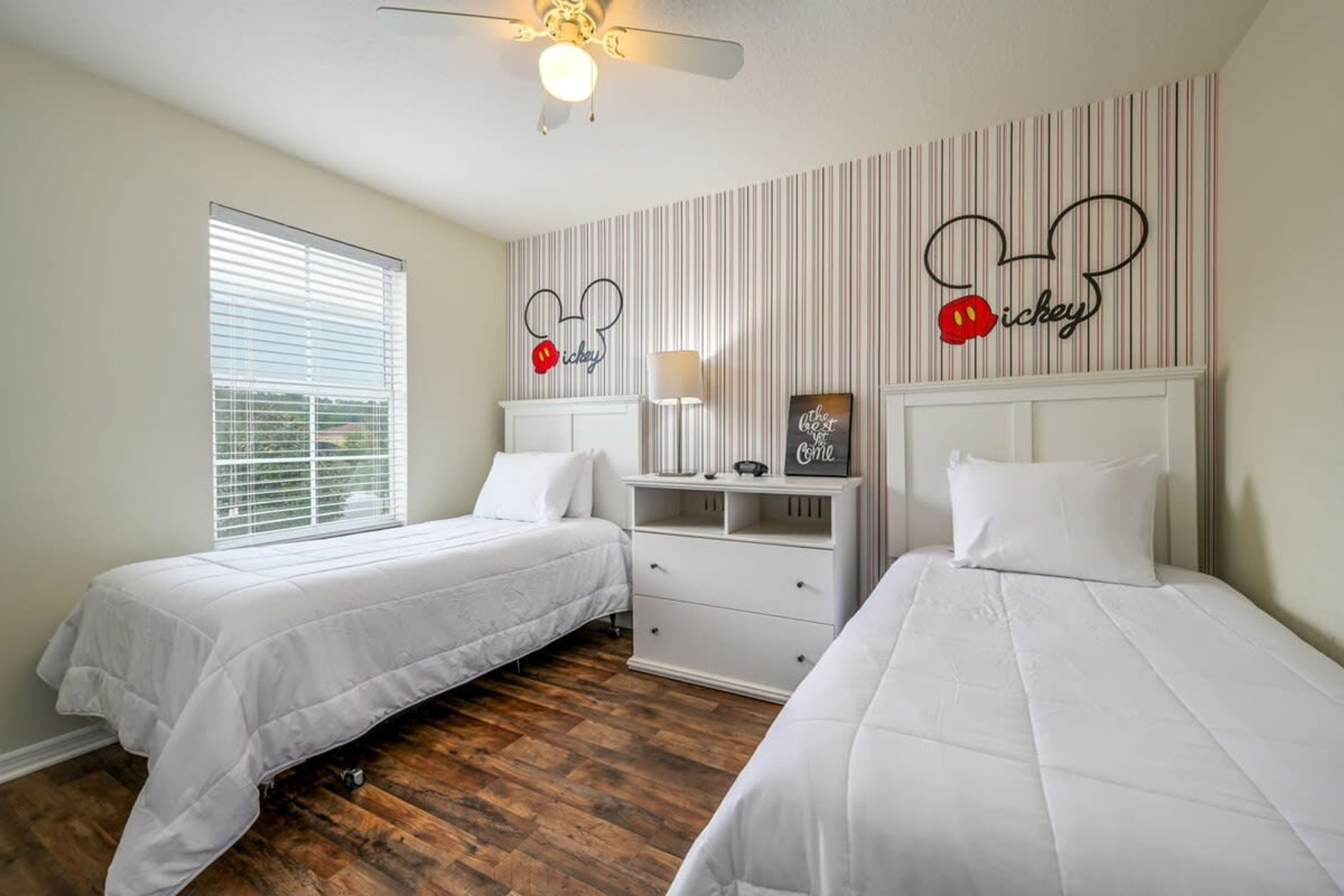 Une chambre avec deux lits, un ventilateur de plafond, une fenêtre avec des stores et une décoration sur le thème de Mickey Mouse.