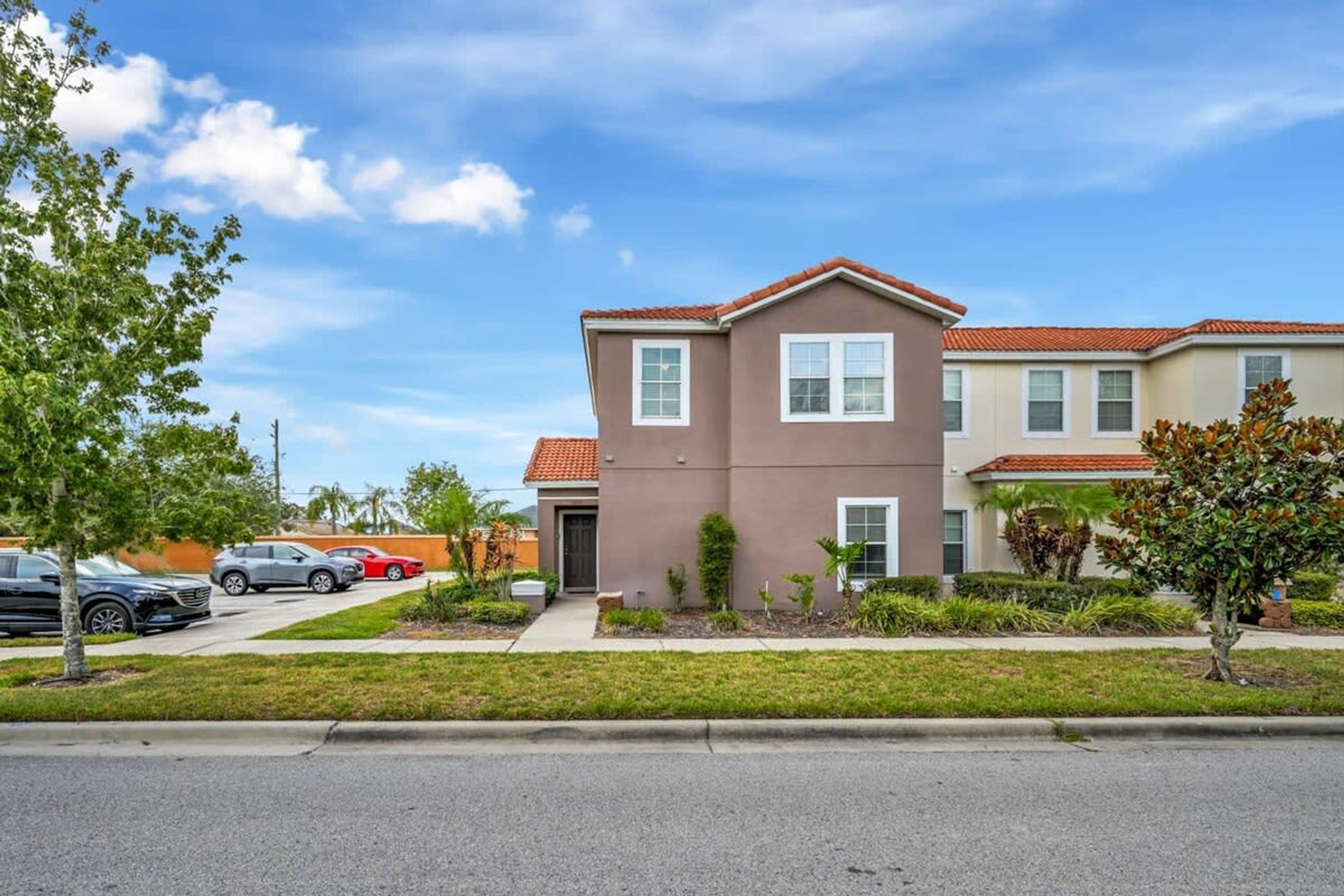 A two-story residential building with a red-tiled roof, a front entrance, and a small front yard with a tree and shrubs.