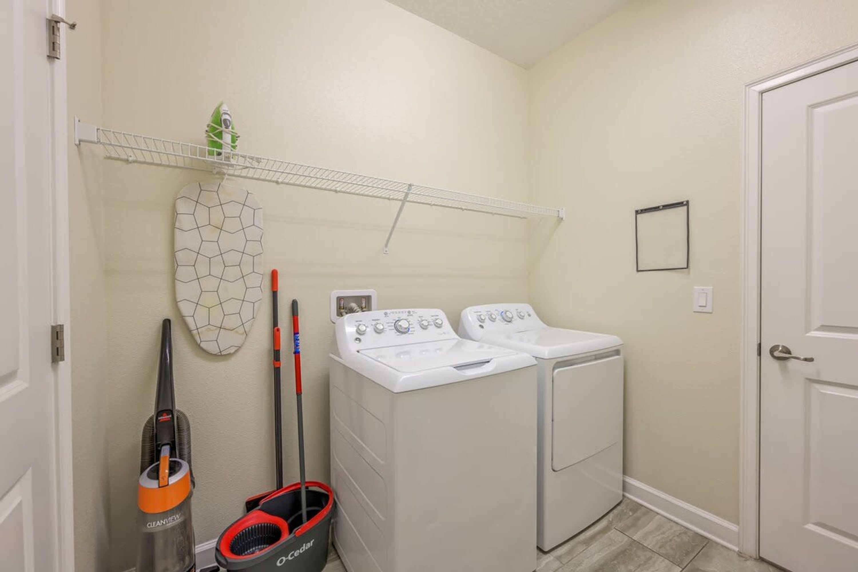 A laundry room with two white front-loading washers and dryers, a broom, a vacuum cleaner, and a shelf with a plant.