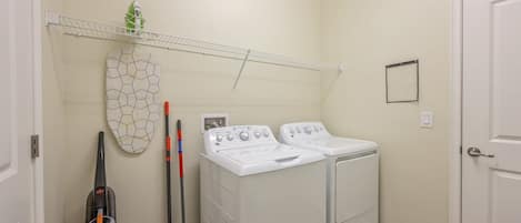 A laundry room with two white front-loading washers and dryers, a broom, a vacuum cleaner, and a shelf with a plant.