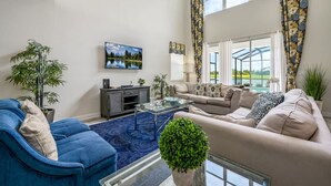 A living room with a blue sofa, beige sofa, and a glass coffee table. There is a painting on the wall and a potted plant in the corner. - Luxury Storey Lake Retreat - Spacious Disney-near (Kissimmee)