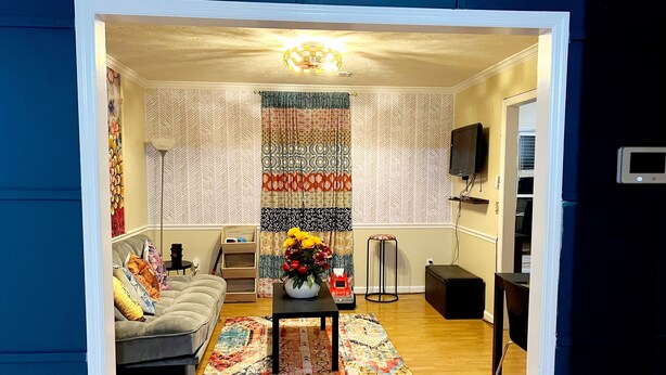 Ein Wohnzimmer mit einer Couch, einem Couchtisch mit Blumenvase, einem gemusterten Teppich, einem Fernseher und einem kleinen Hocker.