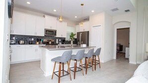 A modern kitchen with white cabinets, a central island with bar stools, stainless steel appliances, and pendant lighting. - Stunning 5BR Home - Your Dream Getaway (Kissimmee)