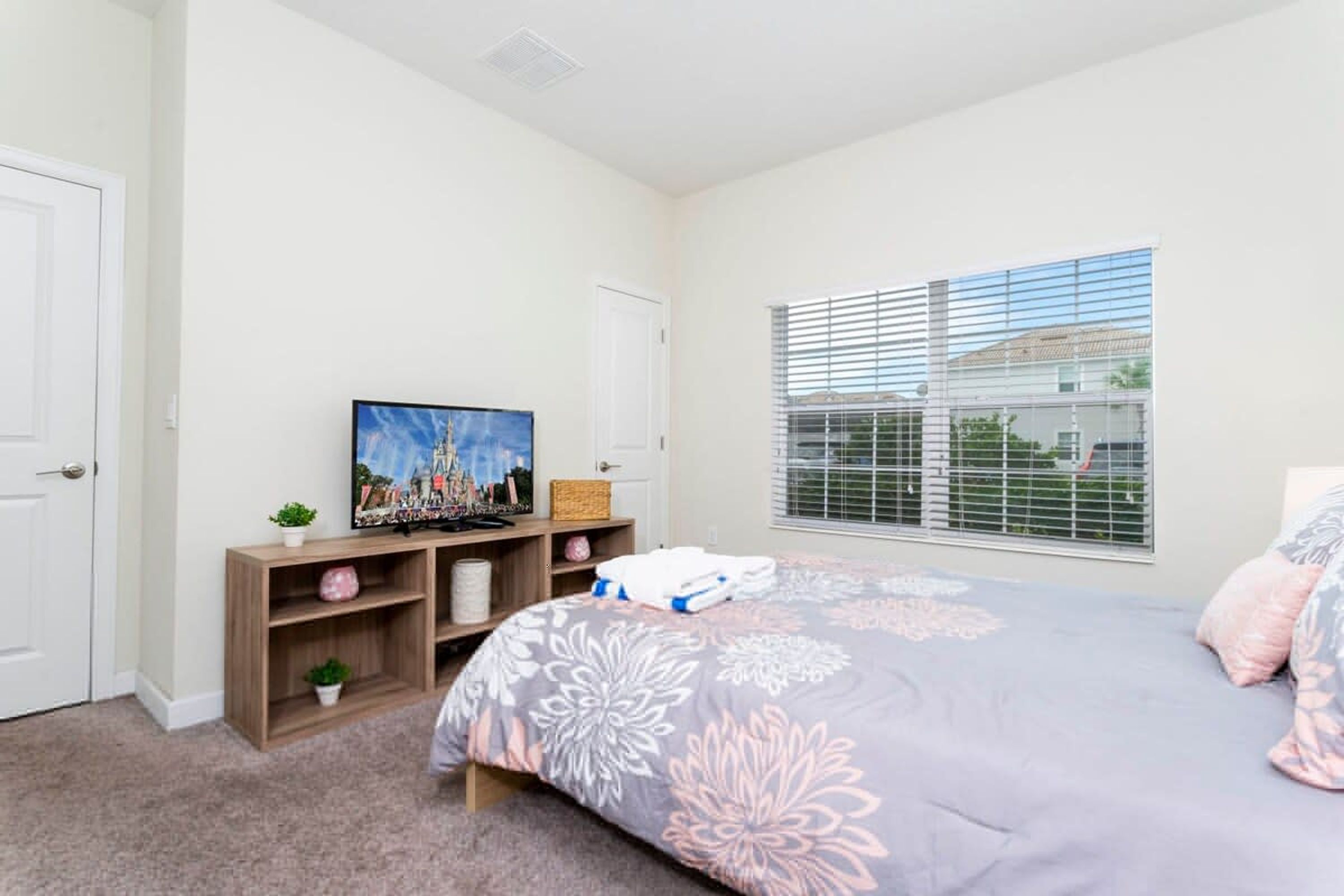 A bedroom with a bed, a television on a stand, a window with blinds, and a door.