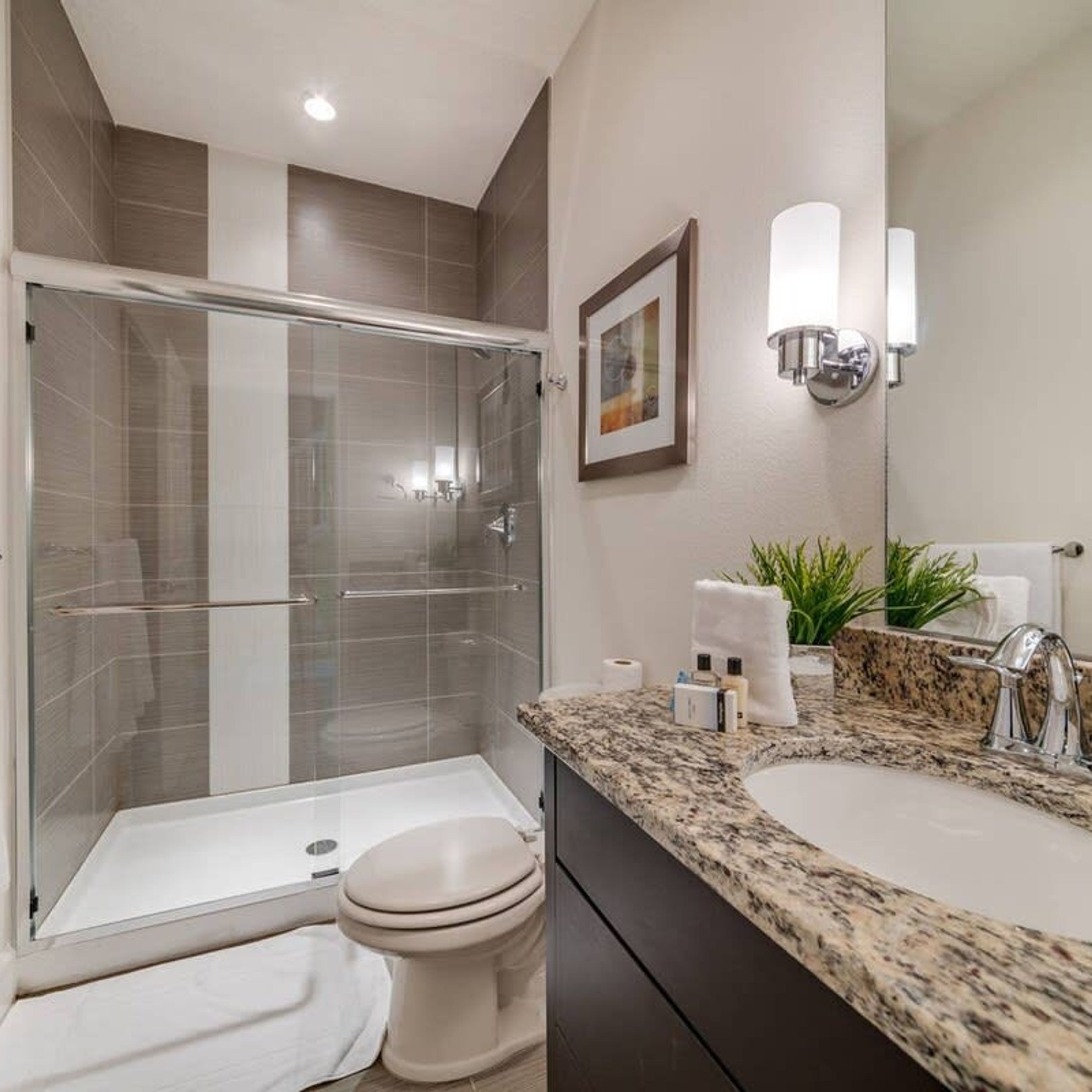 A modern bathroom with a glass-enclosed shower, a toilet, a sink with a granite countertop, and a mirror with wall-mounted lights.