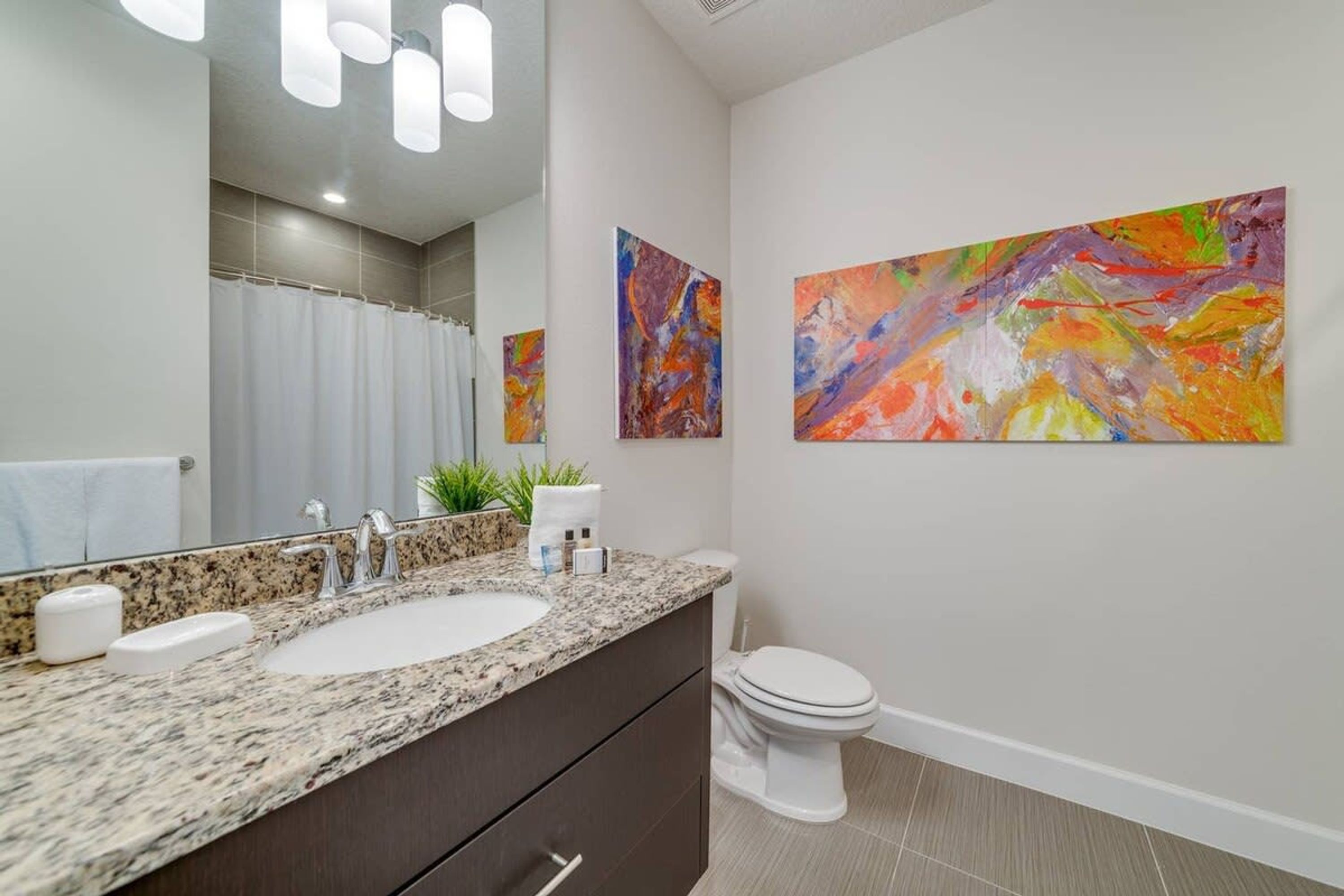 A modern bathroom with a granite countertop, two sinks, a toilet, and a large mirror. There are two abstract paintings on the wall.