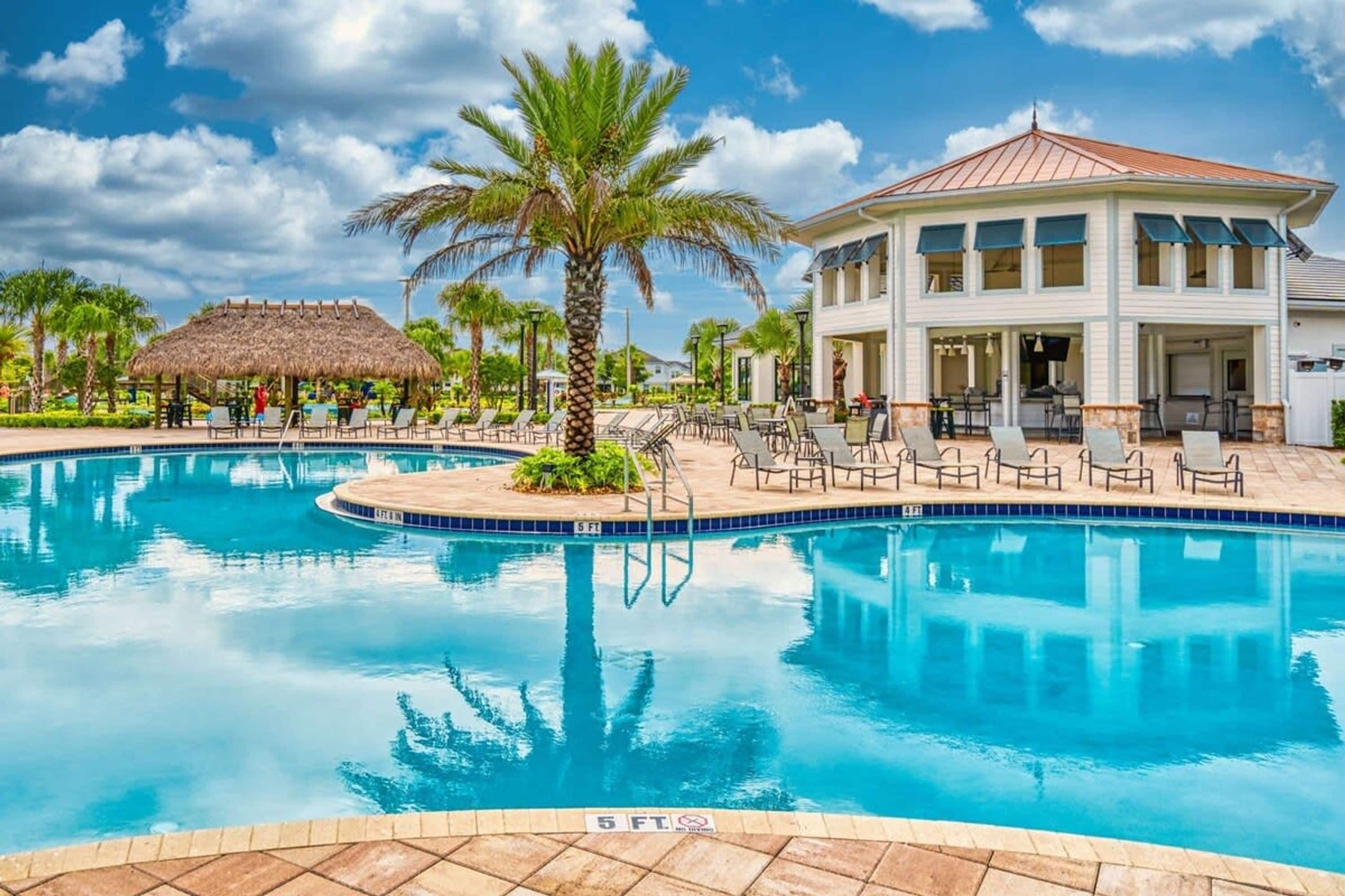 Une piscine avec des chaises longues, un palmier et un bâtiment doté d’un toit de chaume.