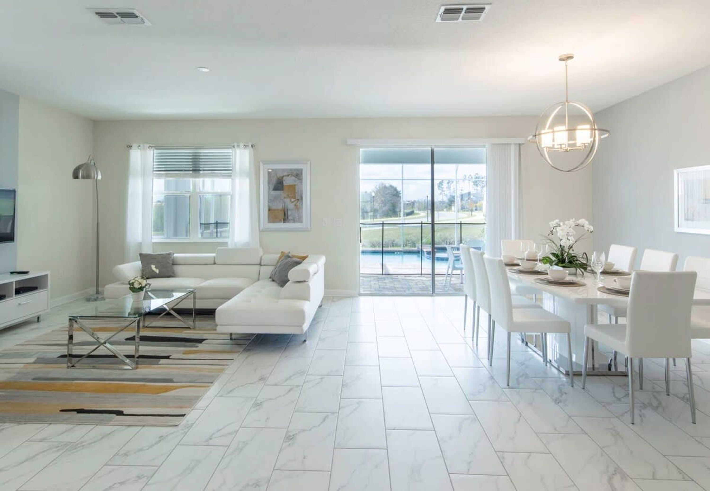 A modern living room with a white sofa, a dining area with a table set for four, and a large window offering a view of a pool.