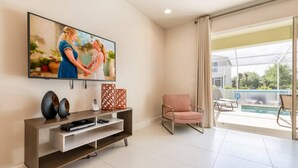 Una sala de estar moderna con un televisor de pantalla plana, un mueble de madera para entretenimiento, un sillón estampado y vistas a una piscina a través de amplias ventanas.