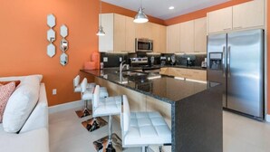 Una cocina moderna con una isla central, electrodomésticos de acero inoxidable y una zona de estar con taburetes blancos.