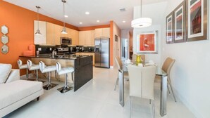 Una cocina moderna con una isla central, taburetes de bar y un área de comedor con mesa y sillas de vidrio.