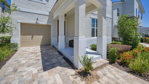 A modern two-story house with a beige garage, a covered porch, and a well-maintained garden with various plants and flowers. - Ultimate Resort Retreat Stunning 6-bedroom Haven (Kissimmee)