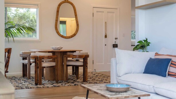 A modern living room with a wooden dining table, white sofa, and a large mirror. - Sunset Cottage - Pet Friendly- Oceanstays (Yamba)