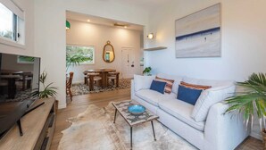 A spacious living room with a sofa, coffee table, and a TV. There is a dining area with a table and chairs, and a large mirror on the wall. - Sunset Cottage - Pet Friendly- Oceanstays (Yamba)